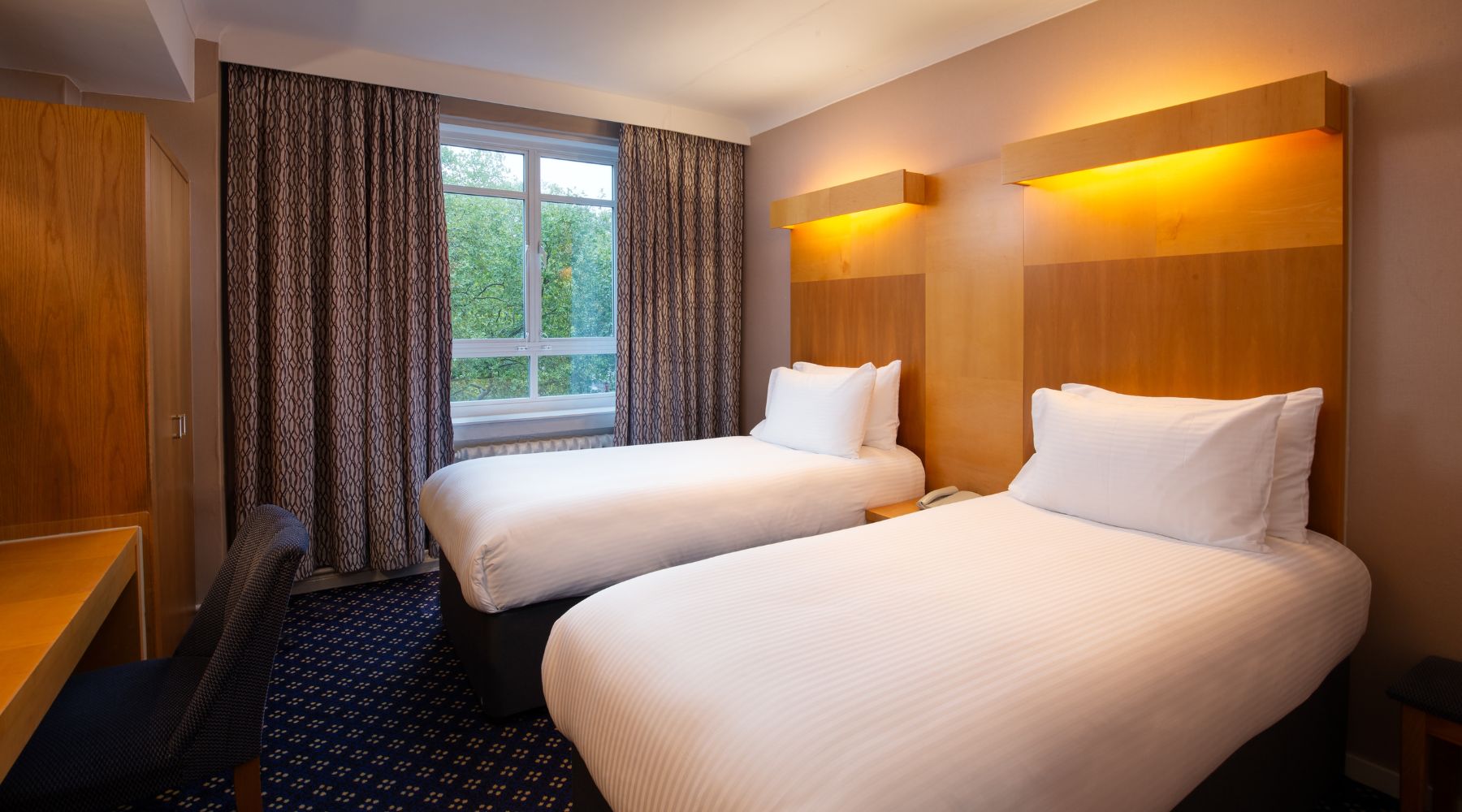 Twin room at Imperial London Hotels with two single beds, warm wood headboards, a desk, and a window facing leafy Bloomsbury.