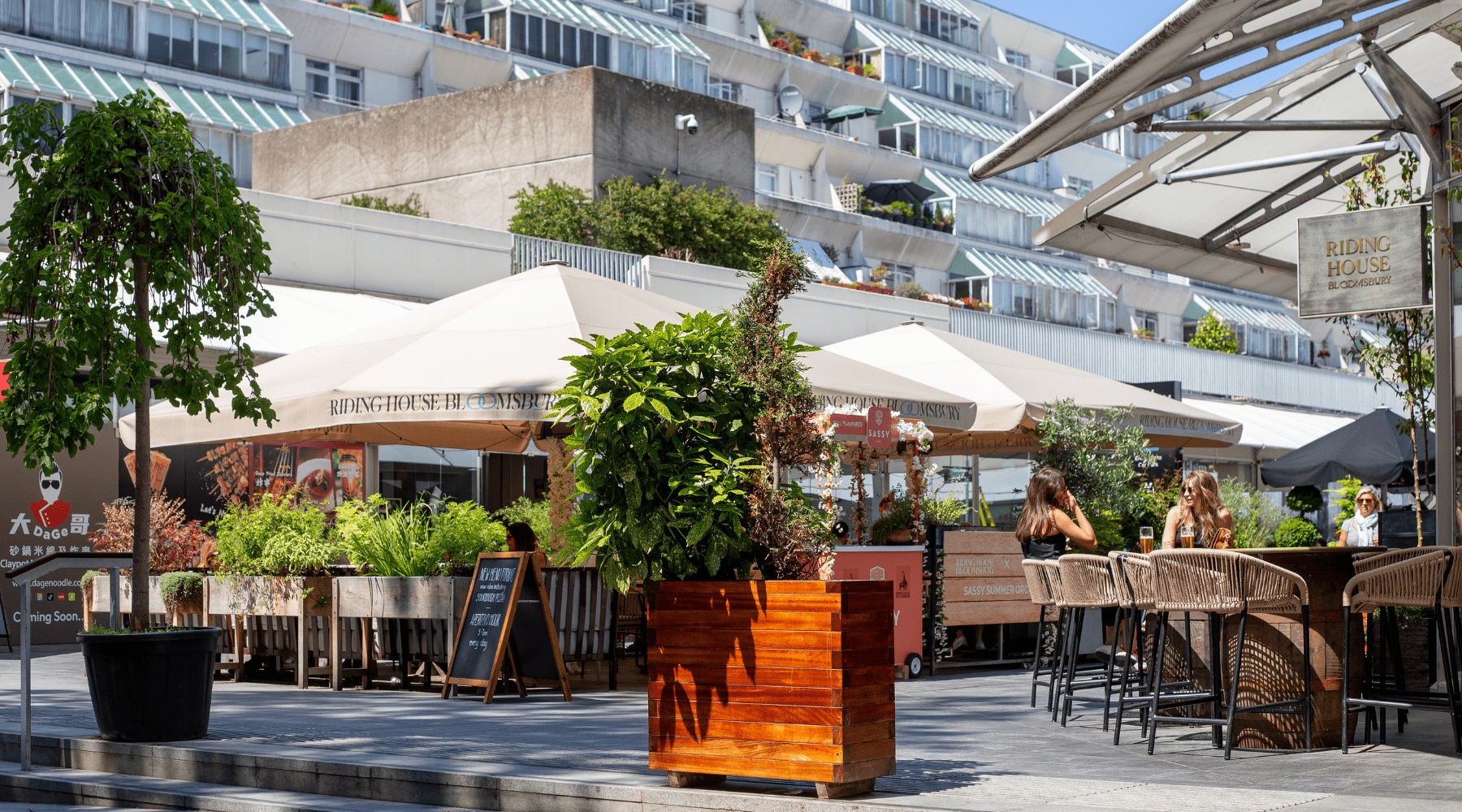 Outdoor café with large umbrellas, greenery in planters, & people dining beside a modern terraced building