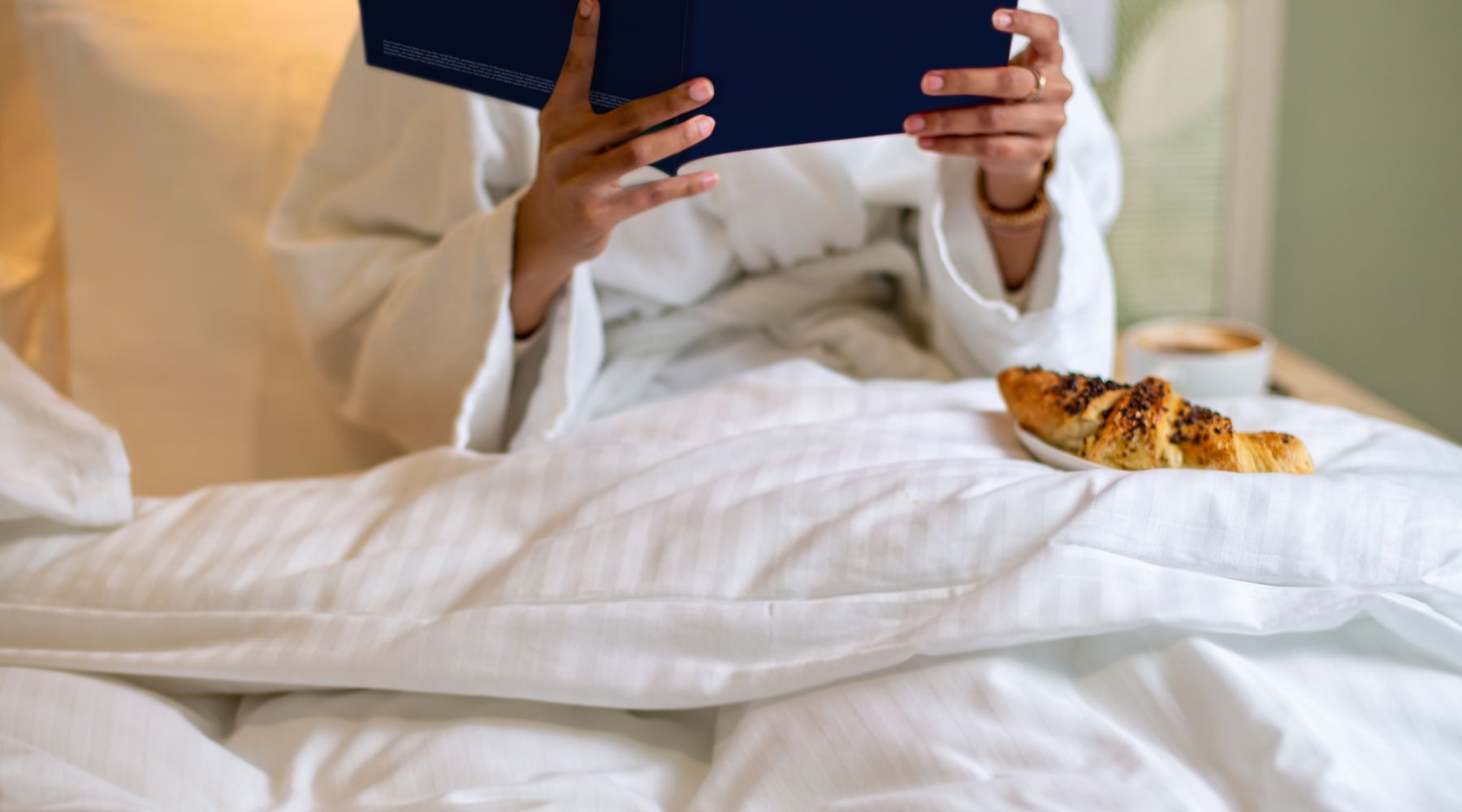 : Guest in a white robe reading a “Bloomsbury London” booklet in an Imperial London Hotels bed, with coffee and a pastry on the duvet.