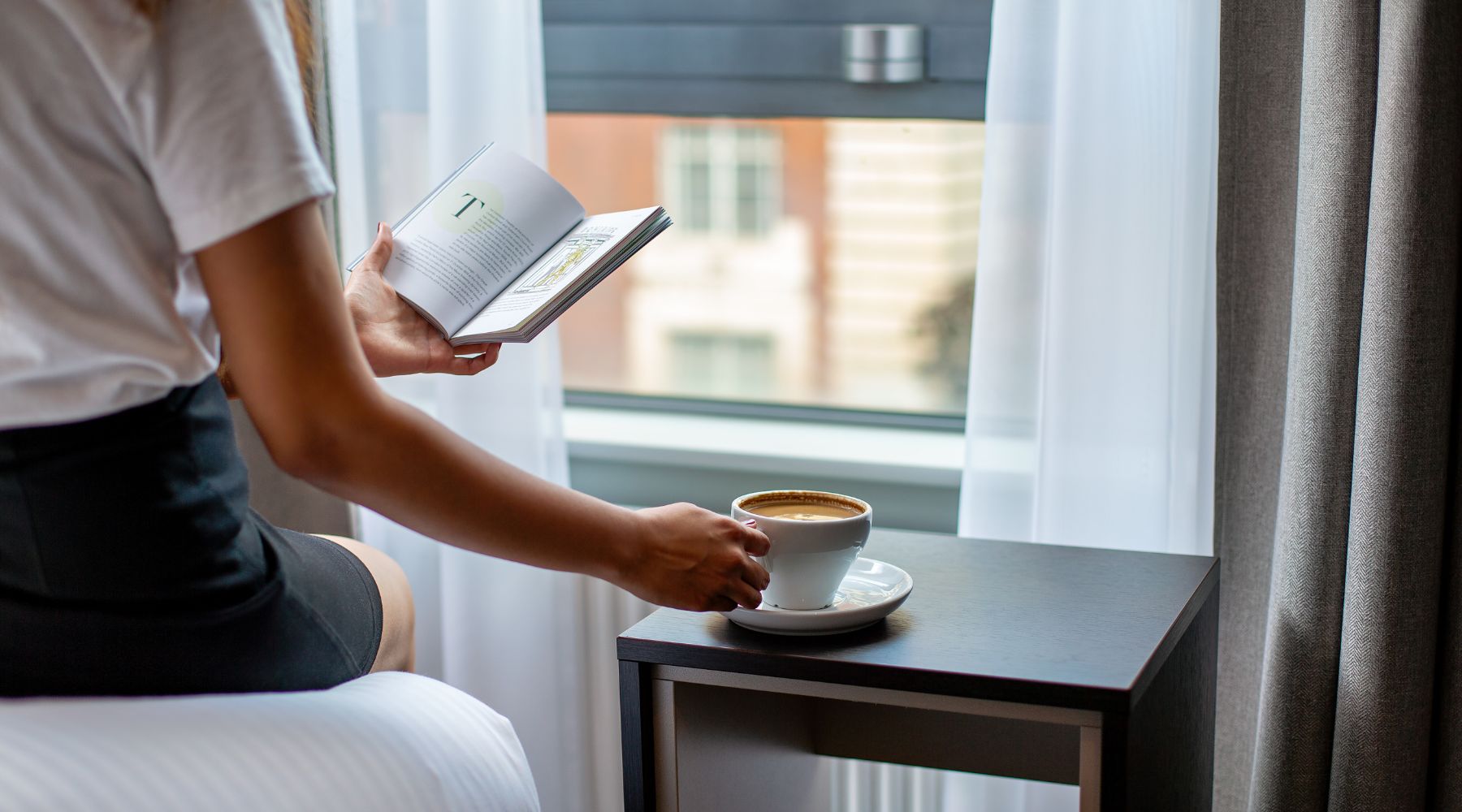 Guest relaxing with a book and coffee by the window in a bright, modern hotel room at Imperial London Hotels, Bloomsbury.