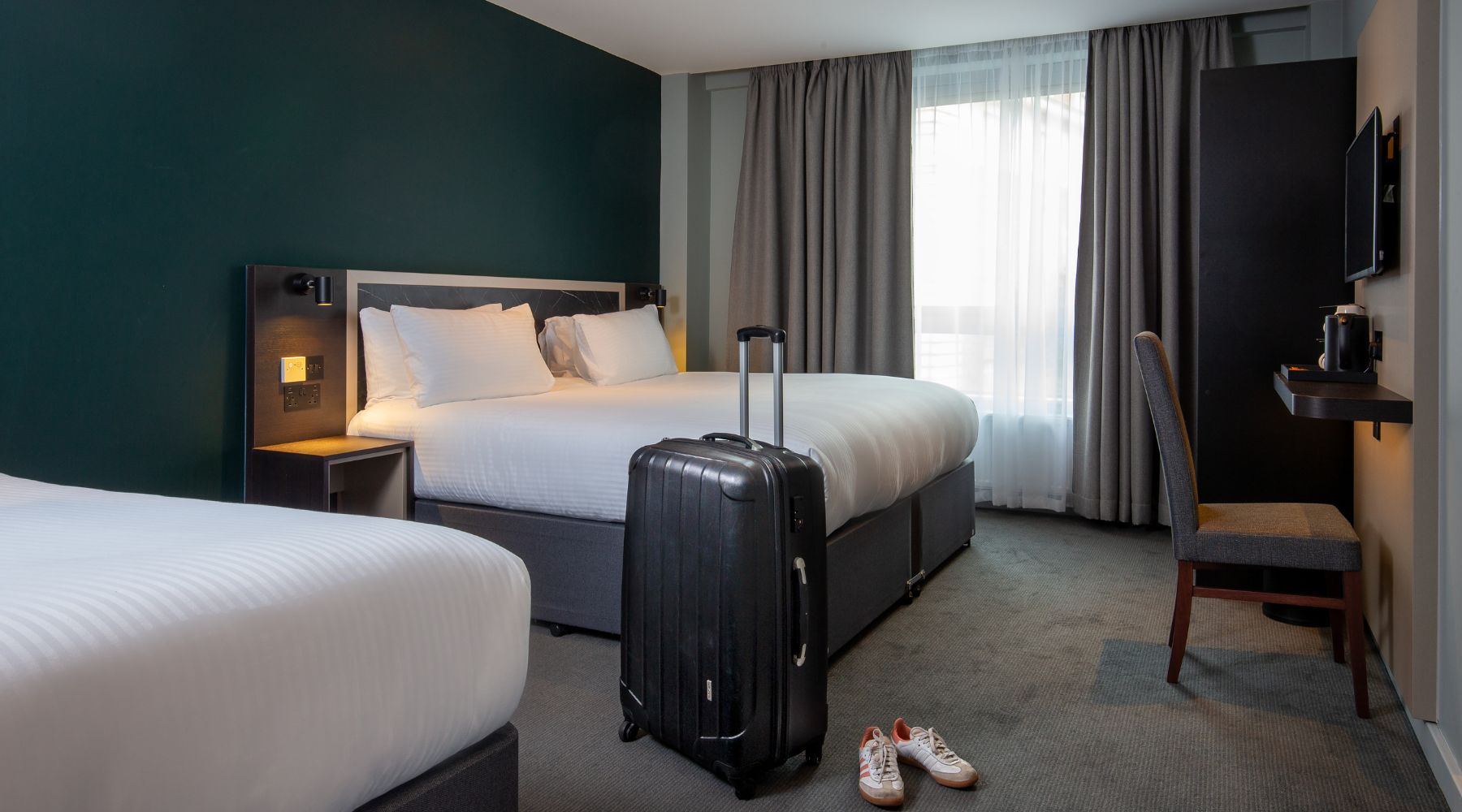 Contemporary twin room with crisp white bedding, teal feature wall, suitcase by the bed, and a desk by the window at Imperial London Hotels.