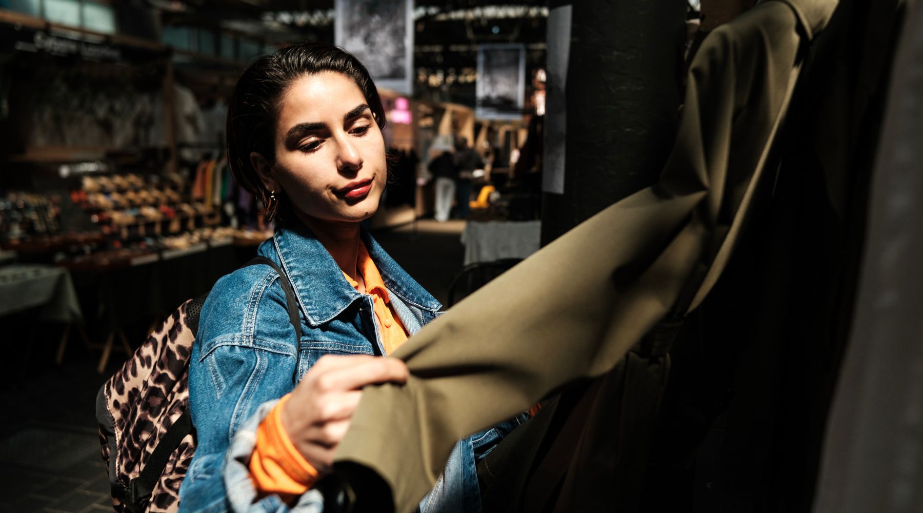 Woman browsing a London market stall, feeling a khaki coat on the rail, wearing a denim jacket and leopard-print backpack.