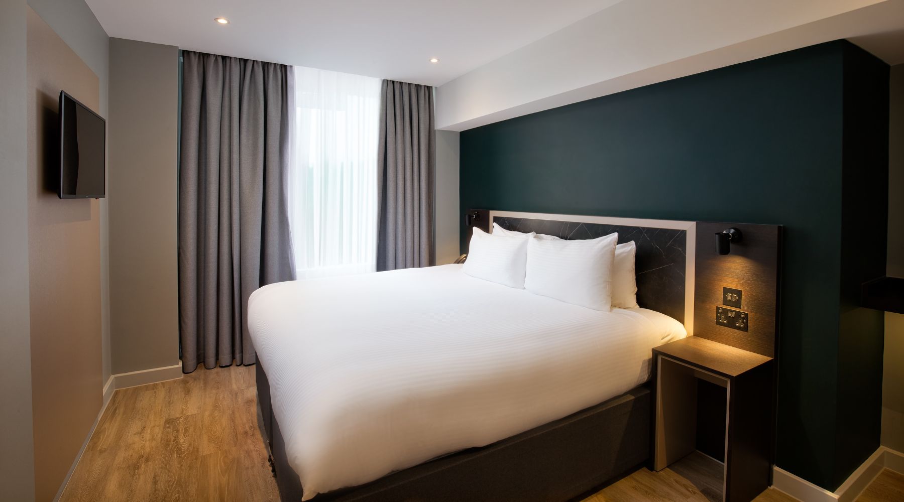 The Royal National Hotel room with a large white bed, dark green accent wall, bedside lighting, curtains, and a mounted TV.