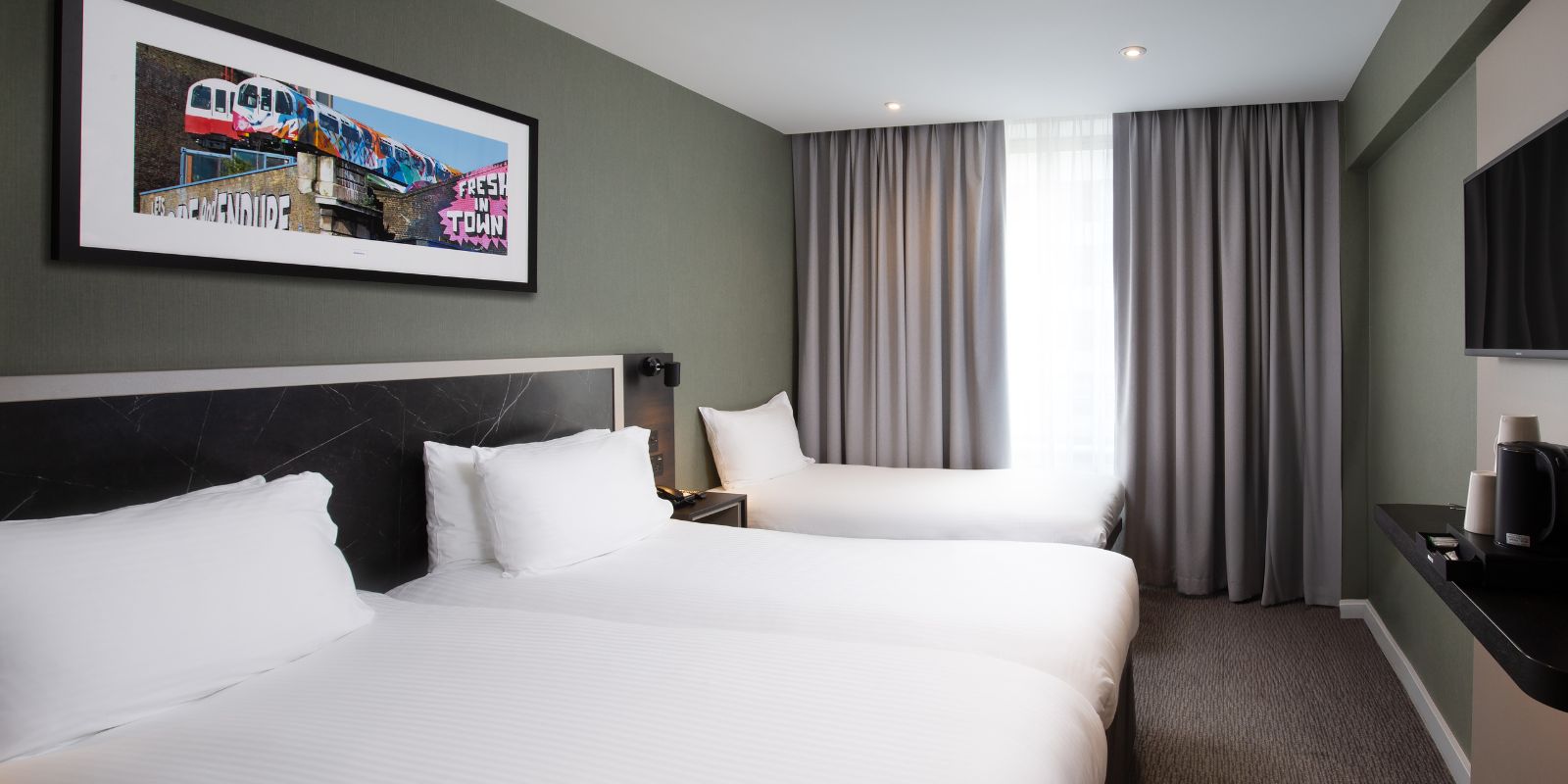 Modern double room at Royal National Hotel: king bed, dark green feature wall, bedside lights and a curtained window.