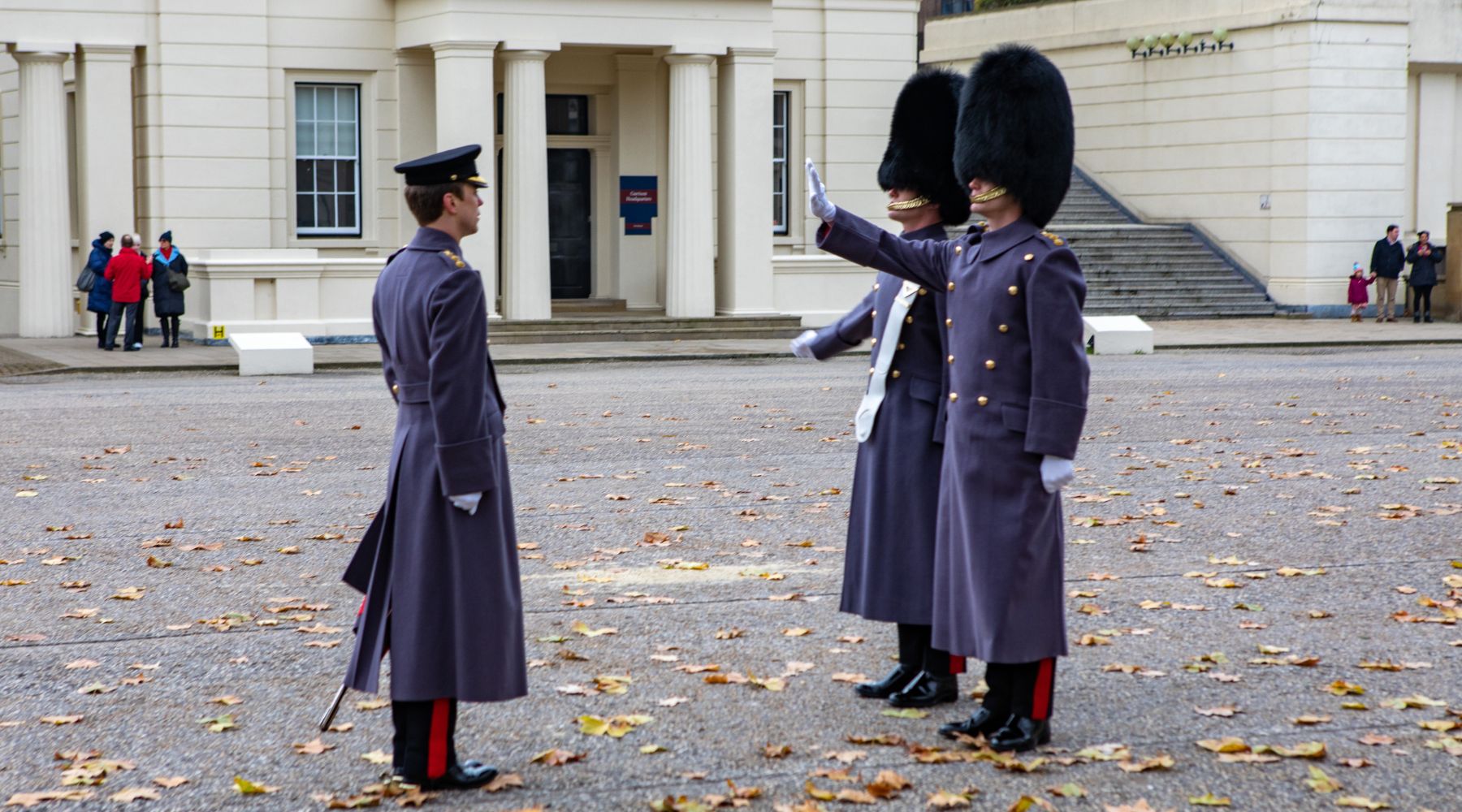  King’s Guard soldiers in bearskin hats and grey greatcoats during a ceremonial drill in London, with fallen autumn leaves underfoot.