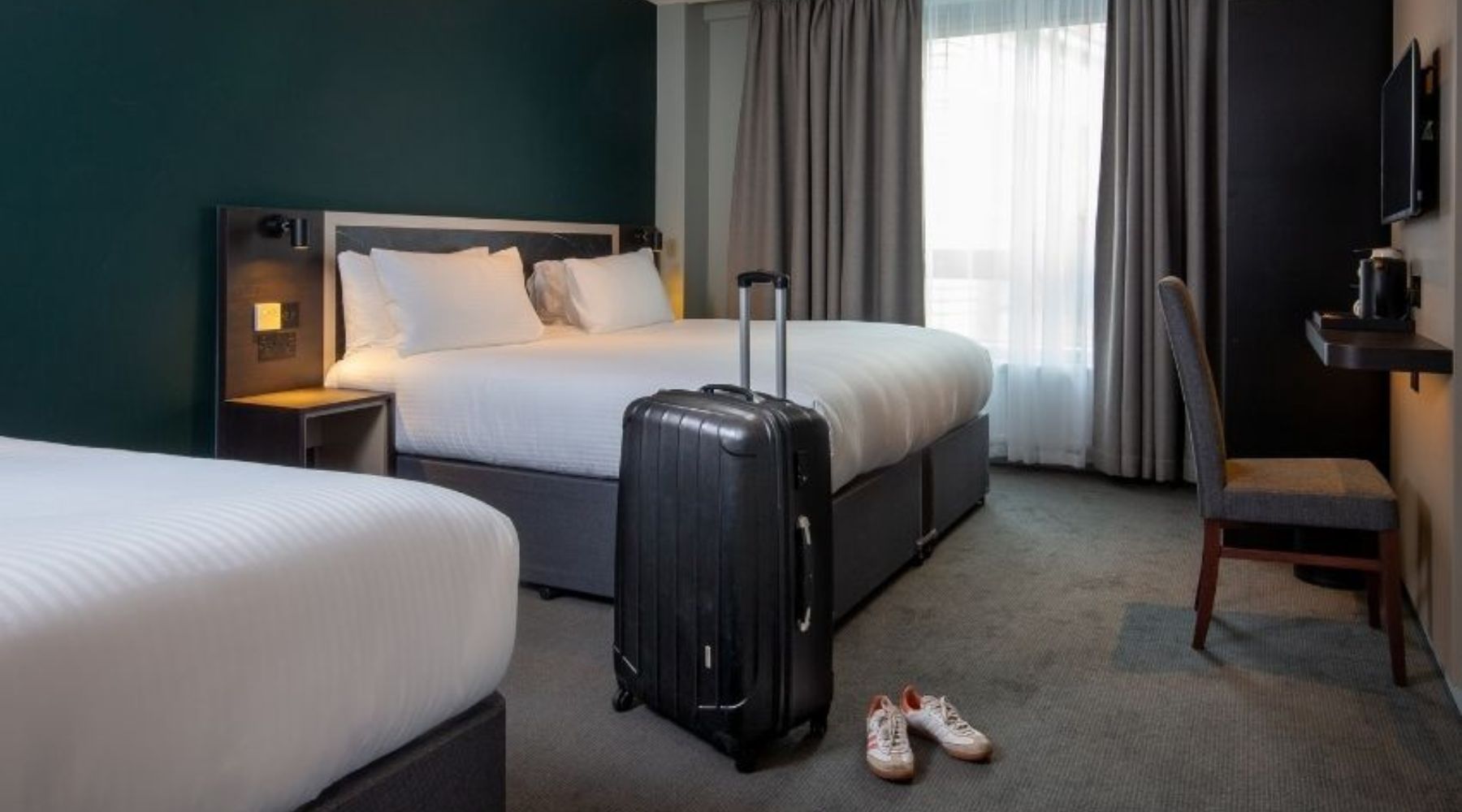Modern hotel room with two beds; upright suitcase by the bed, trainers on carpet, desk and chair, and a window with curtains.