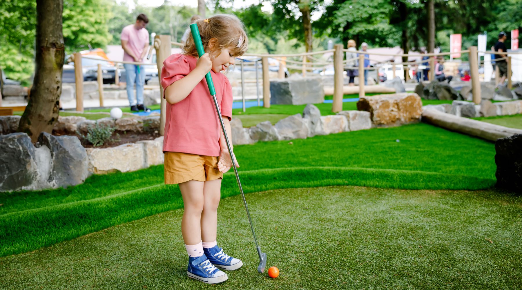 Child in blue trainers lining up a putt on an outdoor mini-golf green, orange ball ready, rocks and landscaping nearby shown.