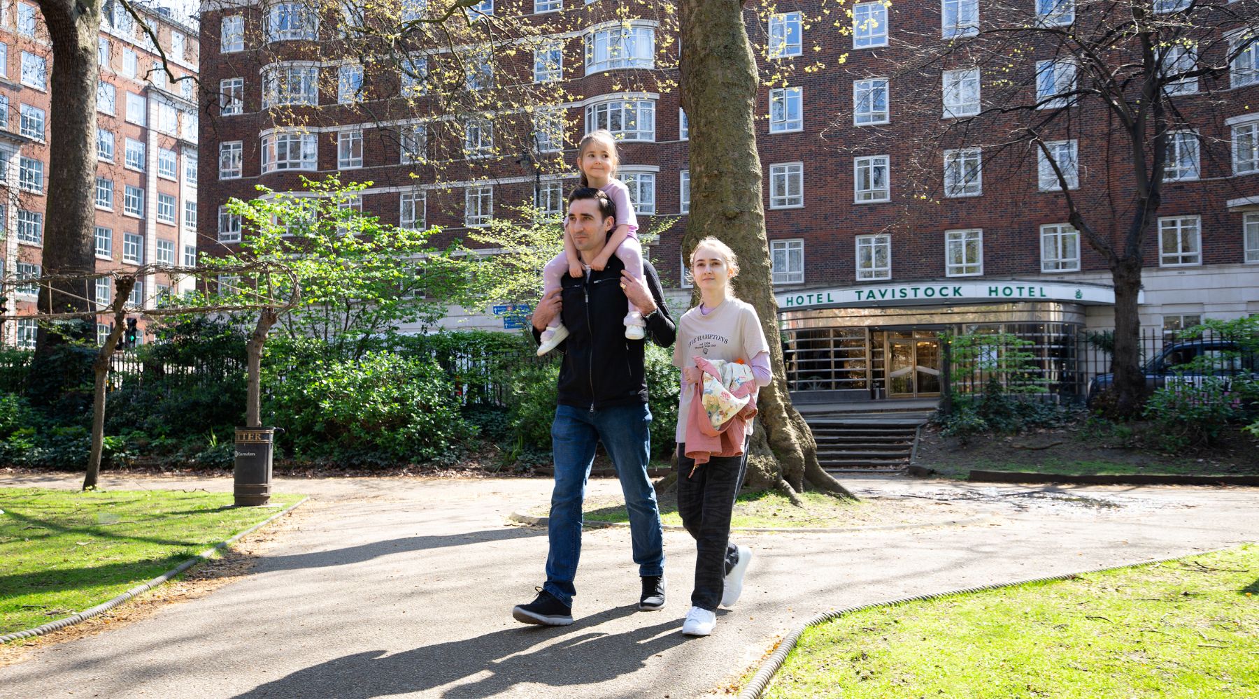 Family walking through a Bloomsbury garden, child on shoulders, with Tavistock Hotel entrance visible in the background.