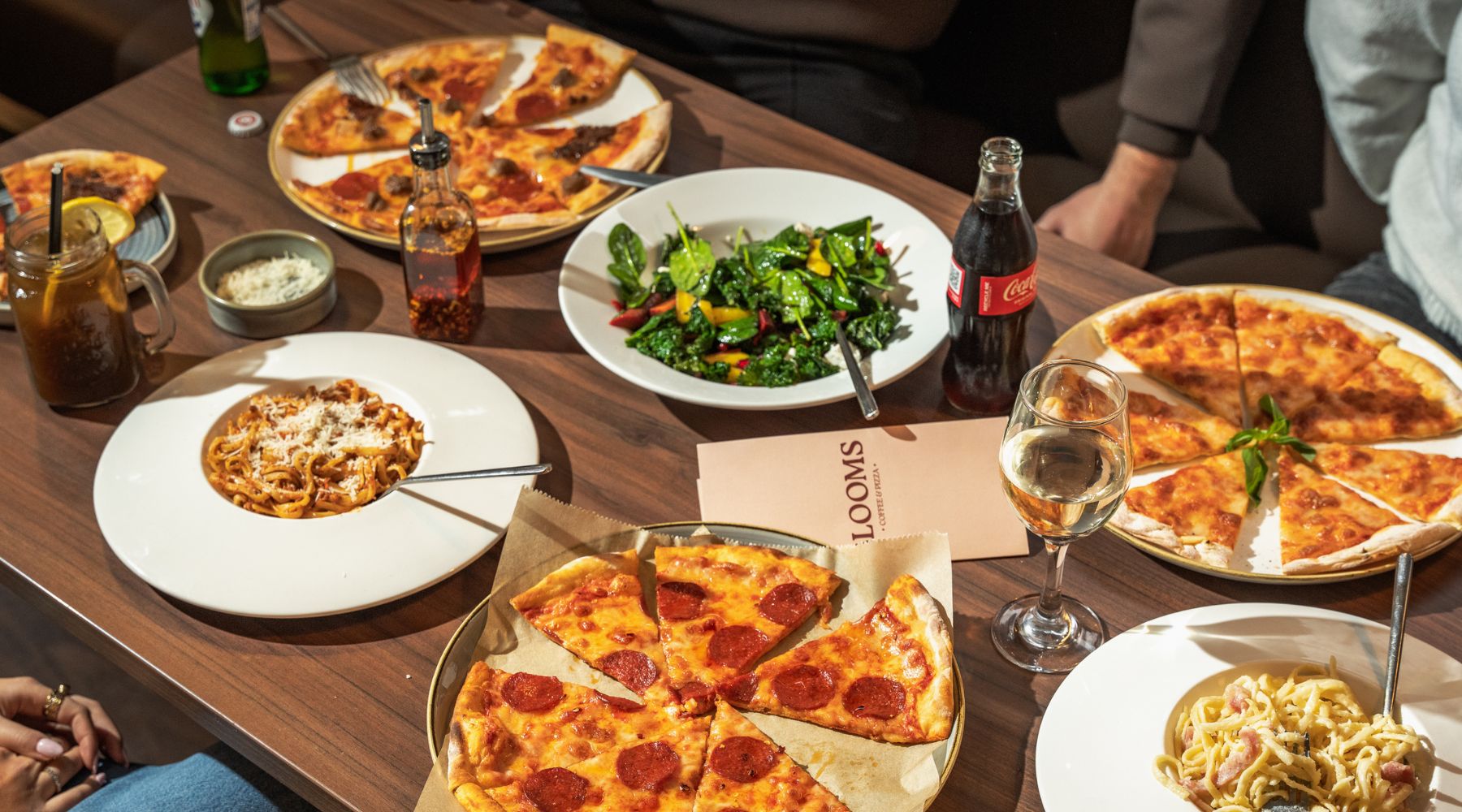 Table spread with pizza, pasta, and salad, served alongside a glass of red wine, a bottle of Coca-Cola, and a Blooms menu placed at the centre.