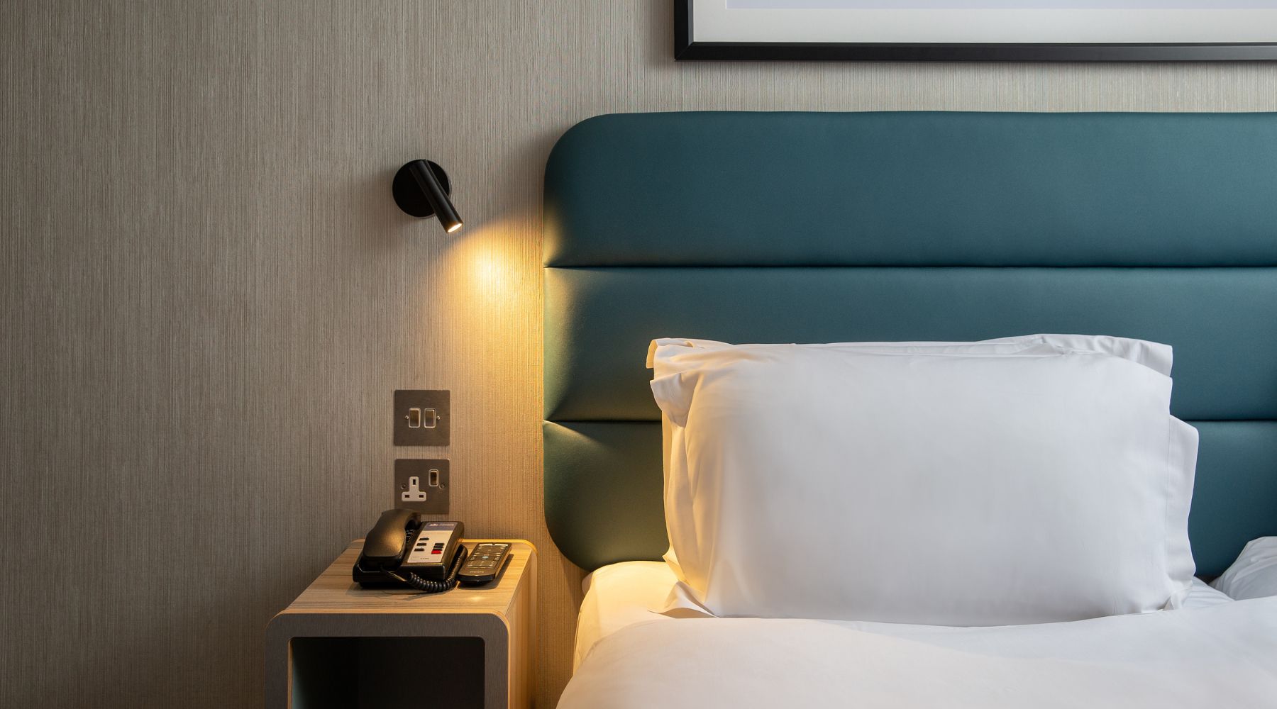 Close-up of a modern hotel bed with teal headboard, bedside lamp, phone, power sockets and USB ports.