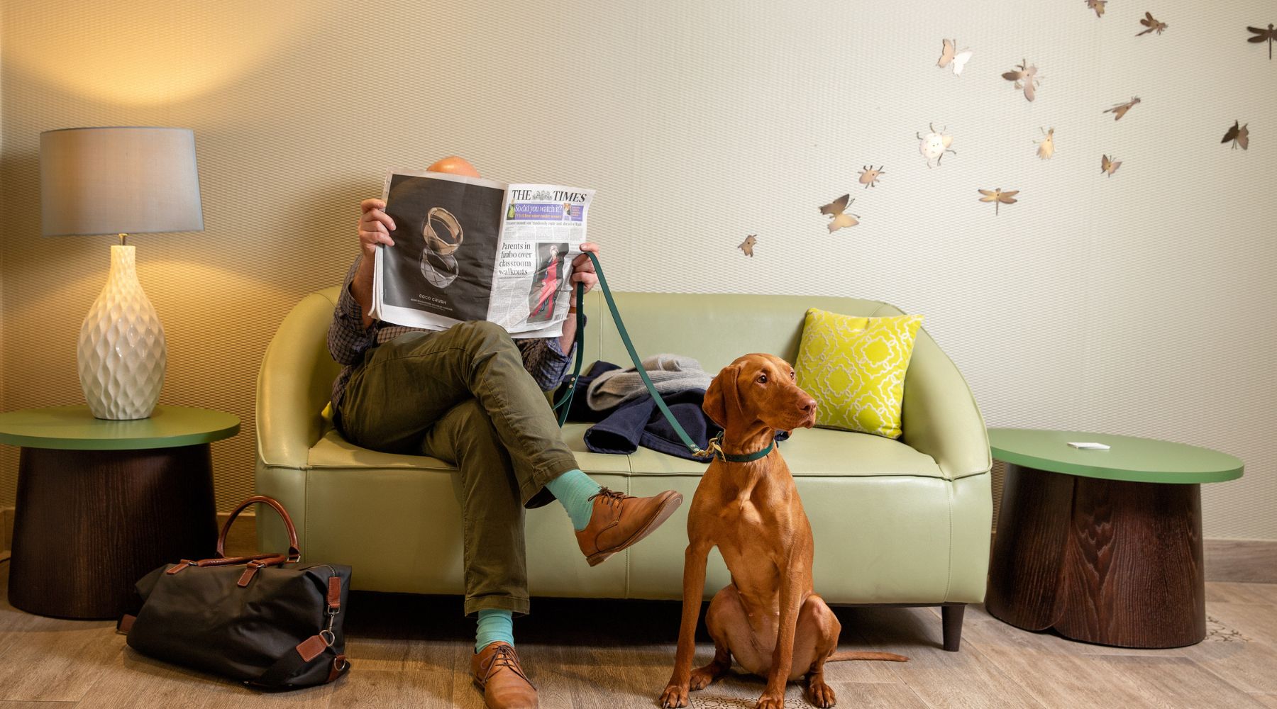 Pet-friendly hotel lobby with a guest reading a newspaper on a green sofa with his dog on a lead, bag and lamps beside.