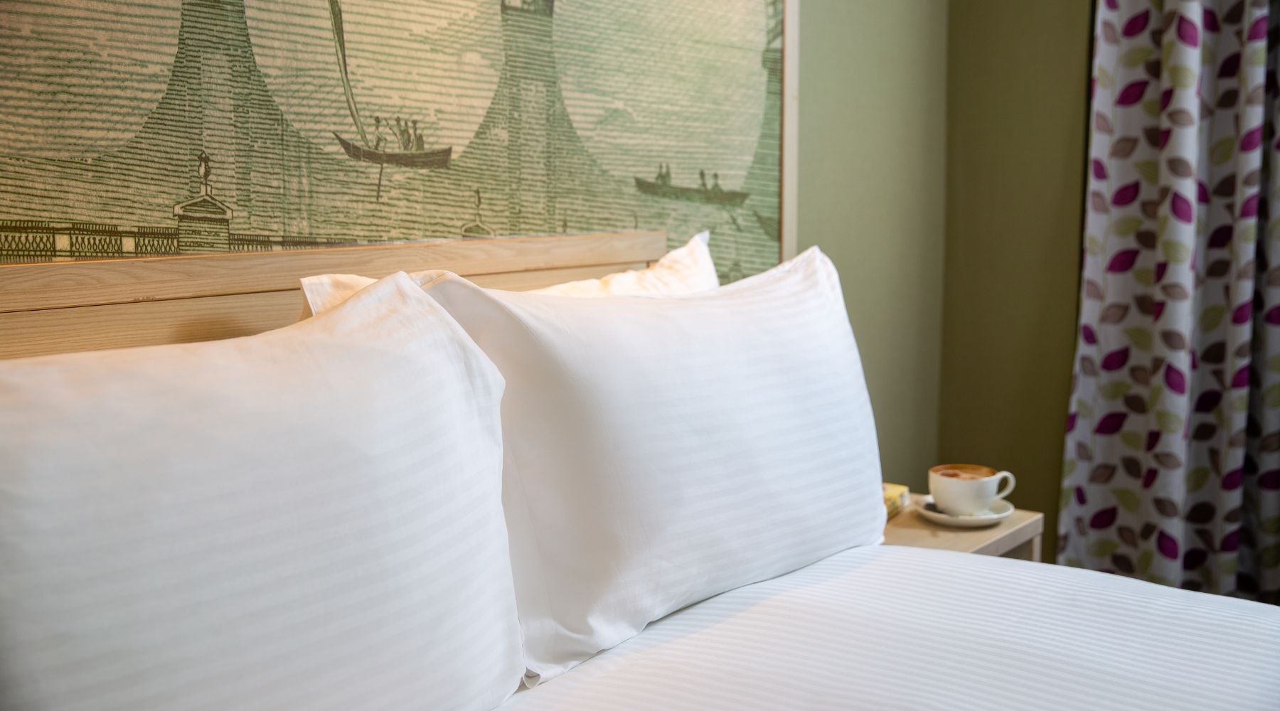 Crisp white bedding with a London mural headboard, patterned curtains and a cup on the bedside table in the President Hotel. 
