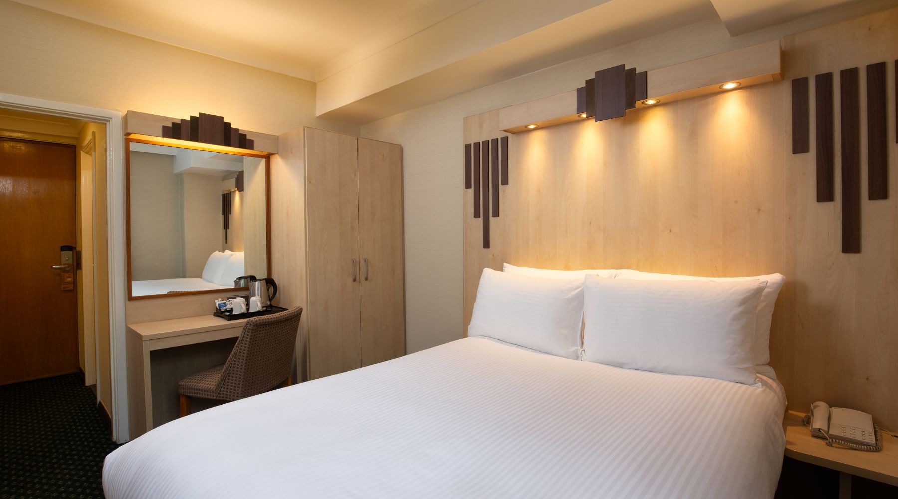 Compact double room with lit wood headboard, white bedding, desk with kettle, wardrobe and mirror in Tavistock Hotel.