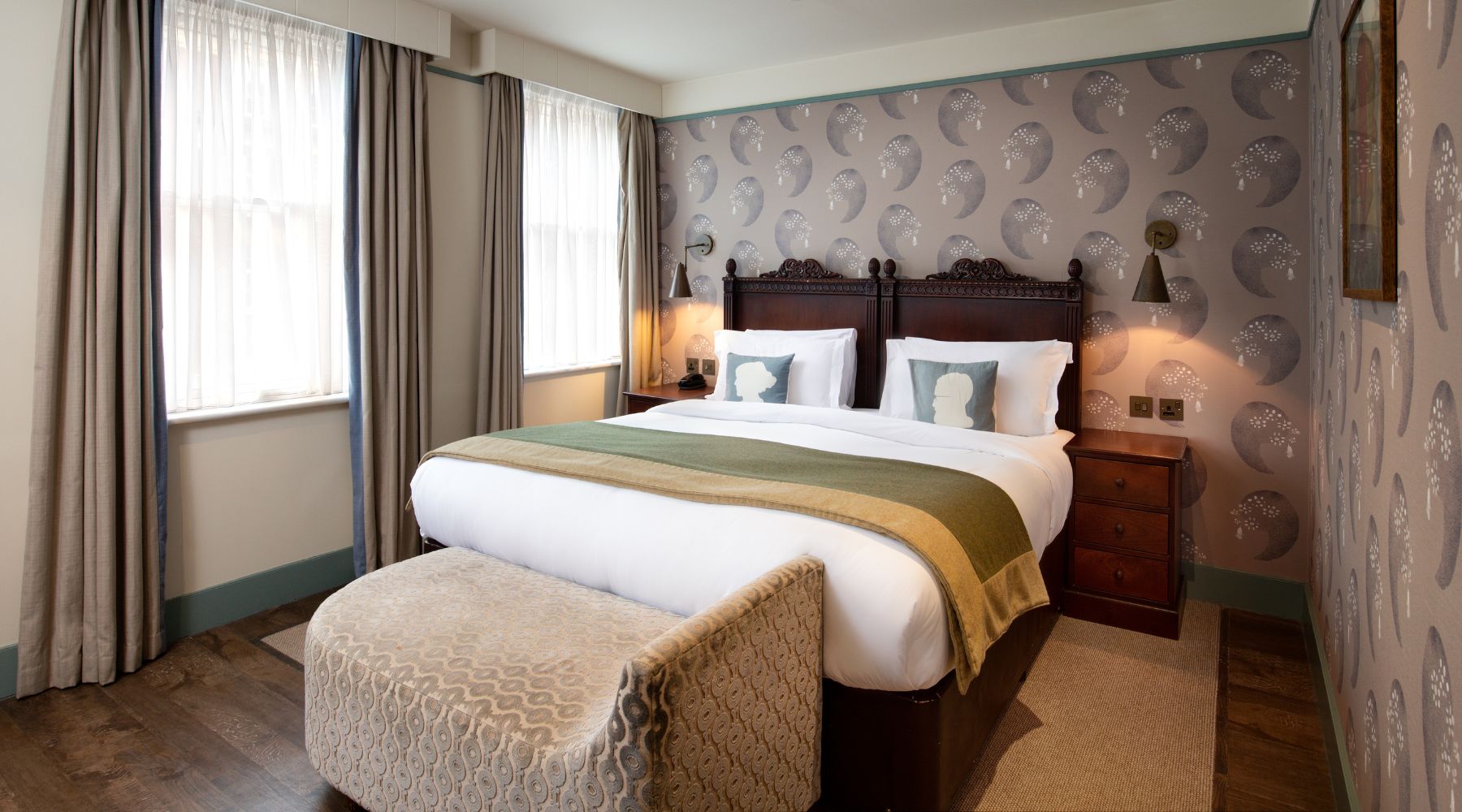 Cosy double room with carved headboard, patterned wallpaper, sash windows, ottoman bench and warm lighting in Morton Hotel. 