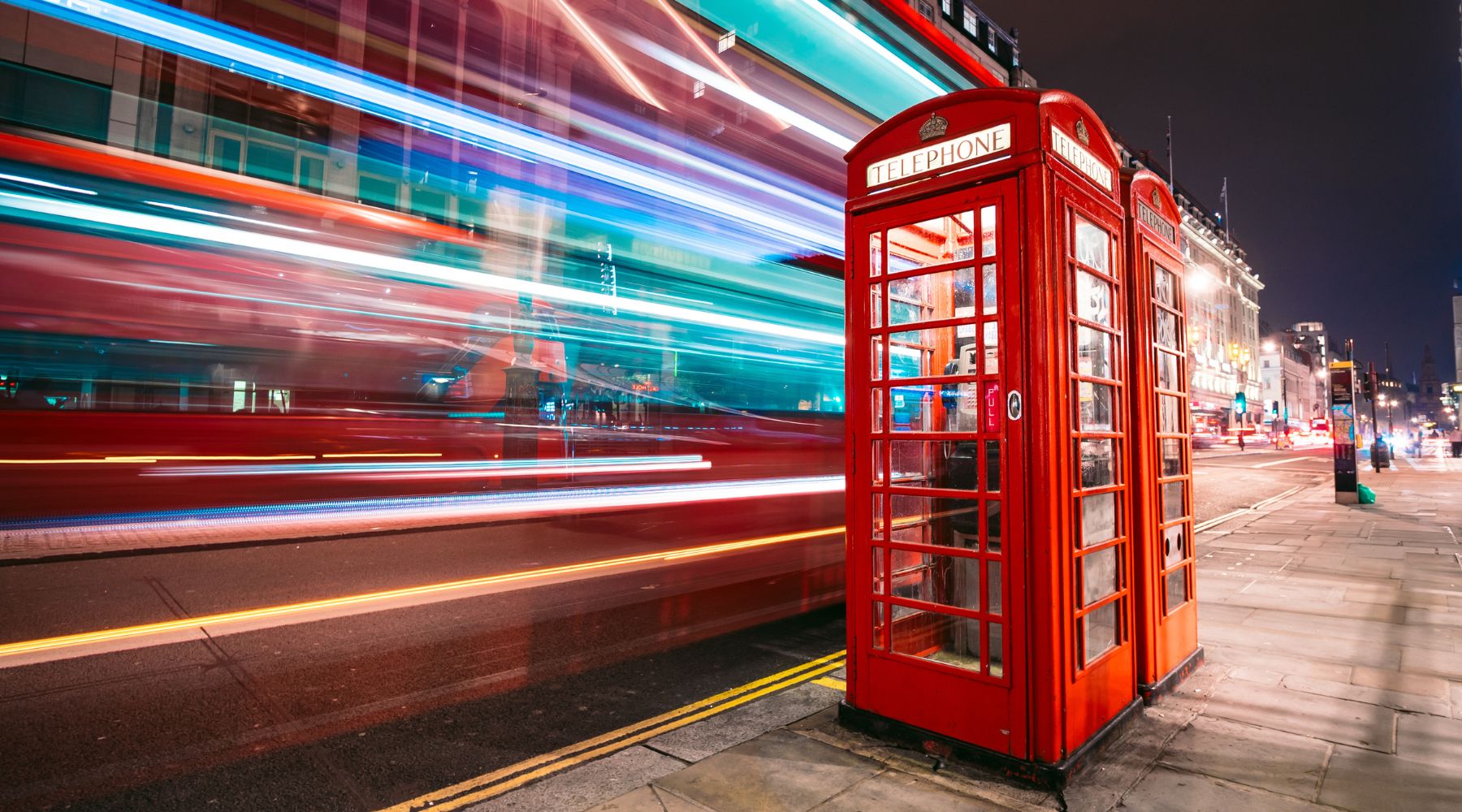  Classic red telephone box with double-decker bus light trails at night on a central London street, capturing energy and style.