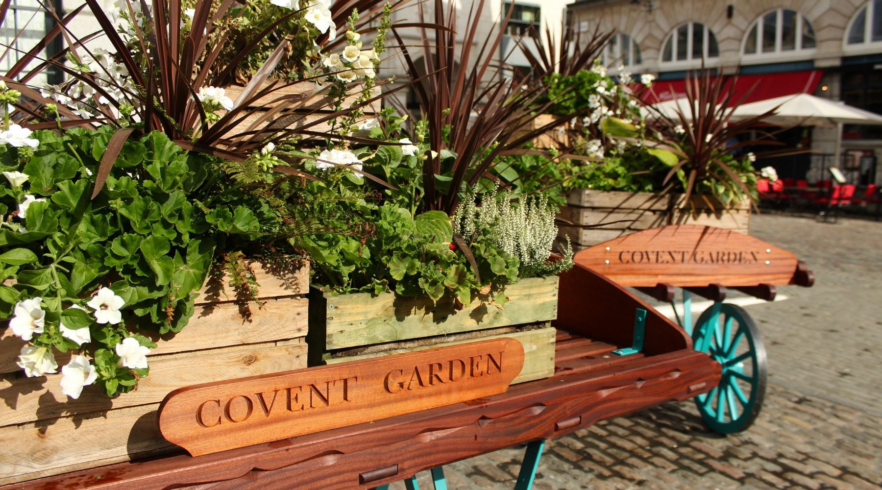 Flower-filled wooden barrows marked ‘Covent Garden’ on cobbles, beside the market with cafés and arches in the background.