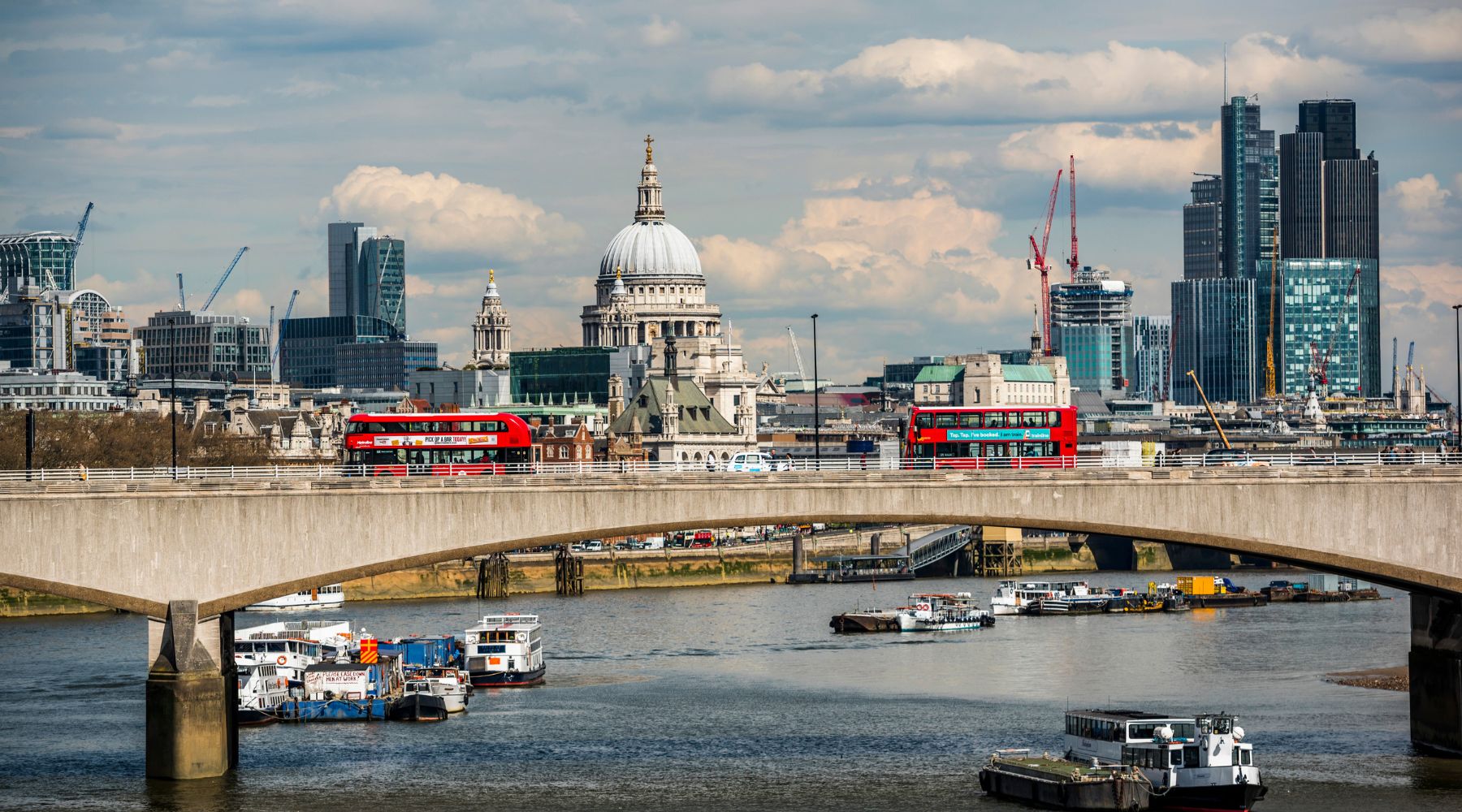 Red double-deckers cross Waterloo Bridge, St Paul’s Cathedral and City skyline behind; boats on the Thames under cloudy sky.