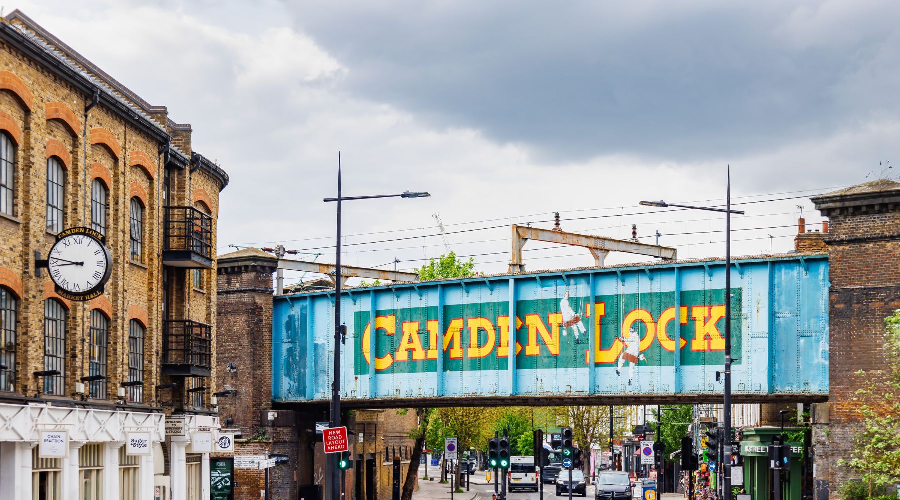Turquoise Camden Lock railway bridge over a road in North London, with brick warehouses, traffic lights and a cloudy sky.