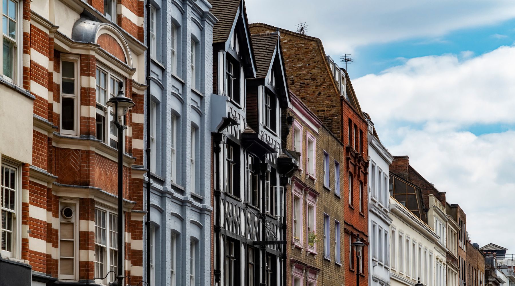 Ornate London townhouses with red brick, pastel façades and Tudor gables line a narrow street beneath a bright, cloudy sky.