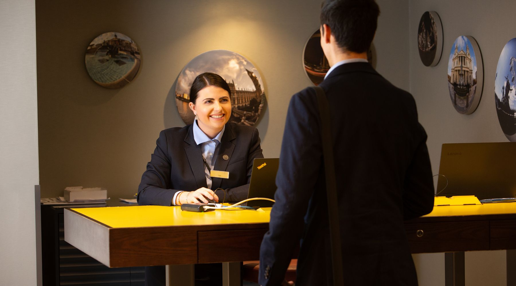 Smiling Imperial London Hotel receptionist at yellow check-in desk greets a guest in a modern lobby, with round London photo wall art behind.