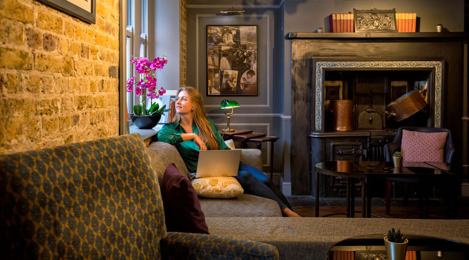 Guest relaxing in a stylish lounge with exposed brick, fireplace and sofa, working on a laptop at Imperial London Hotels.