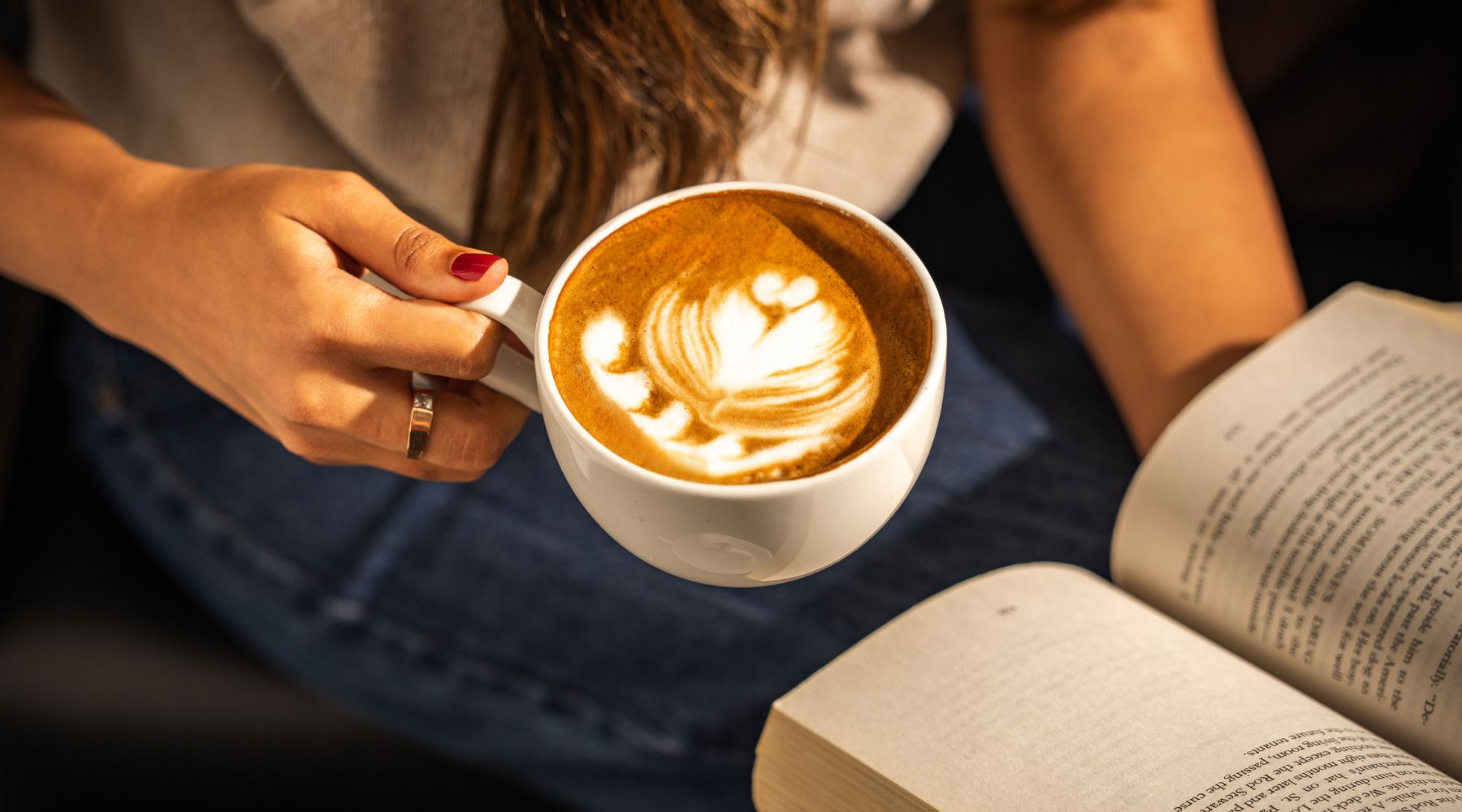 Close-up of a guest holding a latte with leaf art beside an open book, relaxing in a cosy London café setting.
