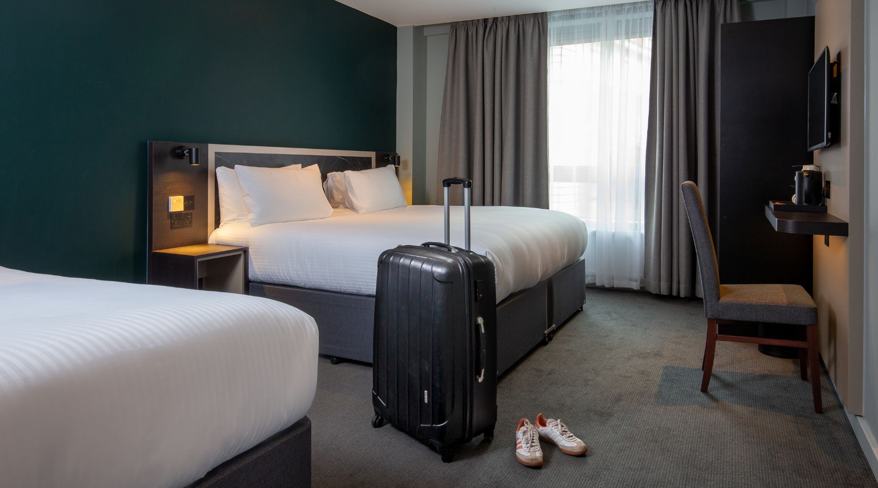 Modern twin hotel room with two single beds, teal feature wall, suitcase by the bed and desk by the window at Imperial London Hotels.